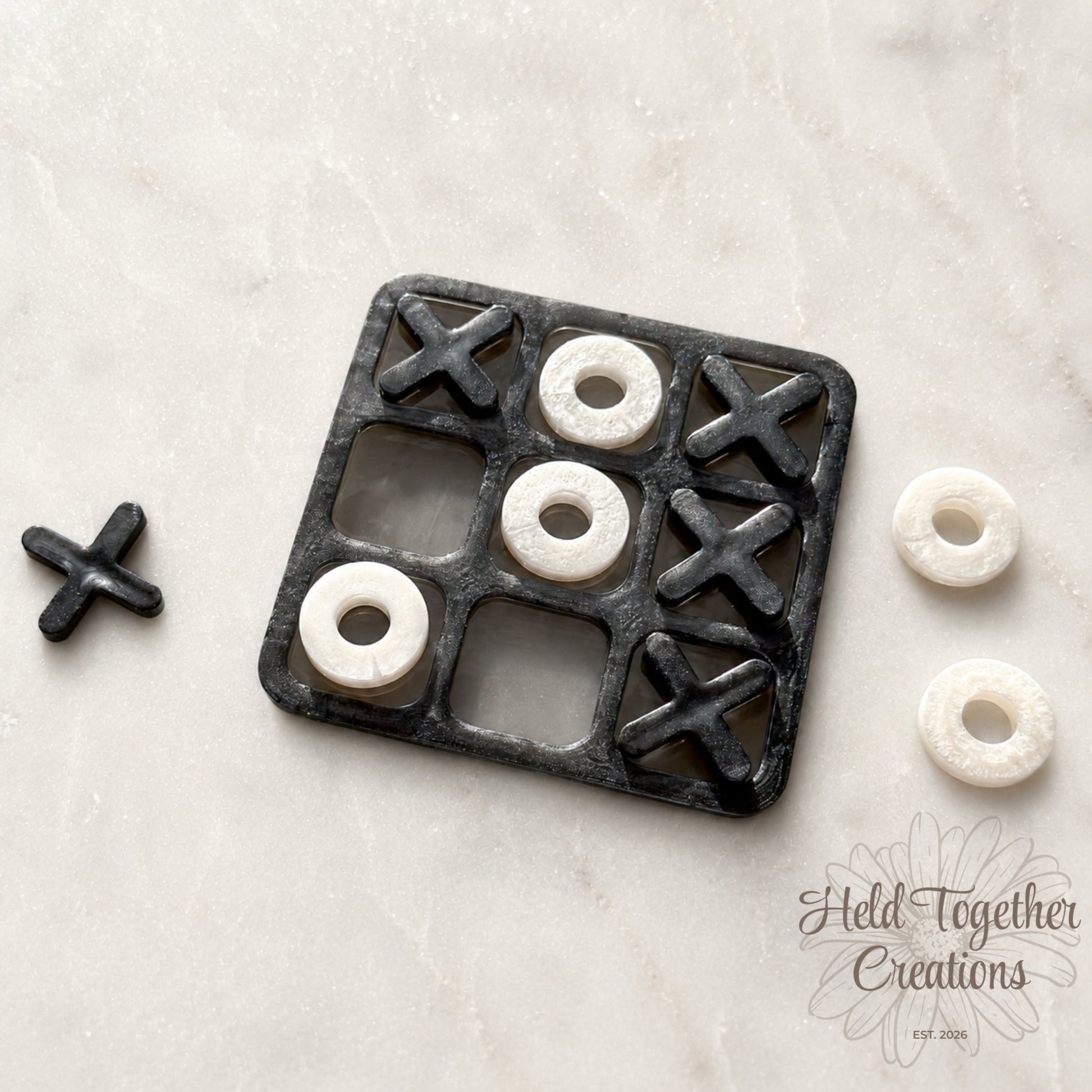 Handmade monochrome resin tic-tac-toe game with black board, black X pieces and white O pieces on a soft neutral background.