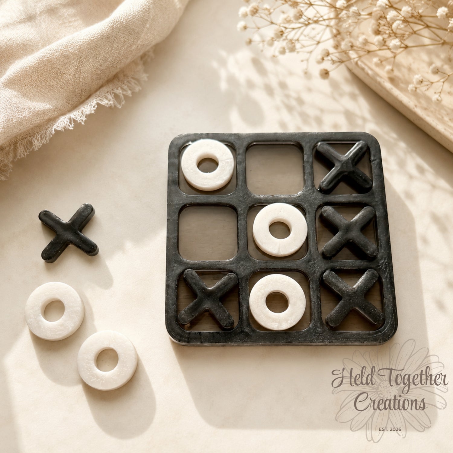 Handmade monochrome resin tic-tac-toe game with black board, black X pieces and white O pieces on a soft neutral background.