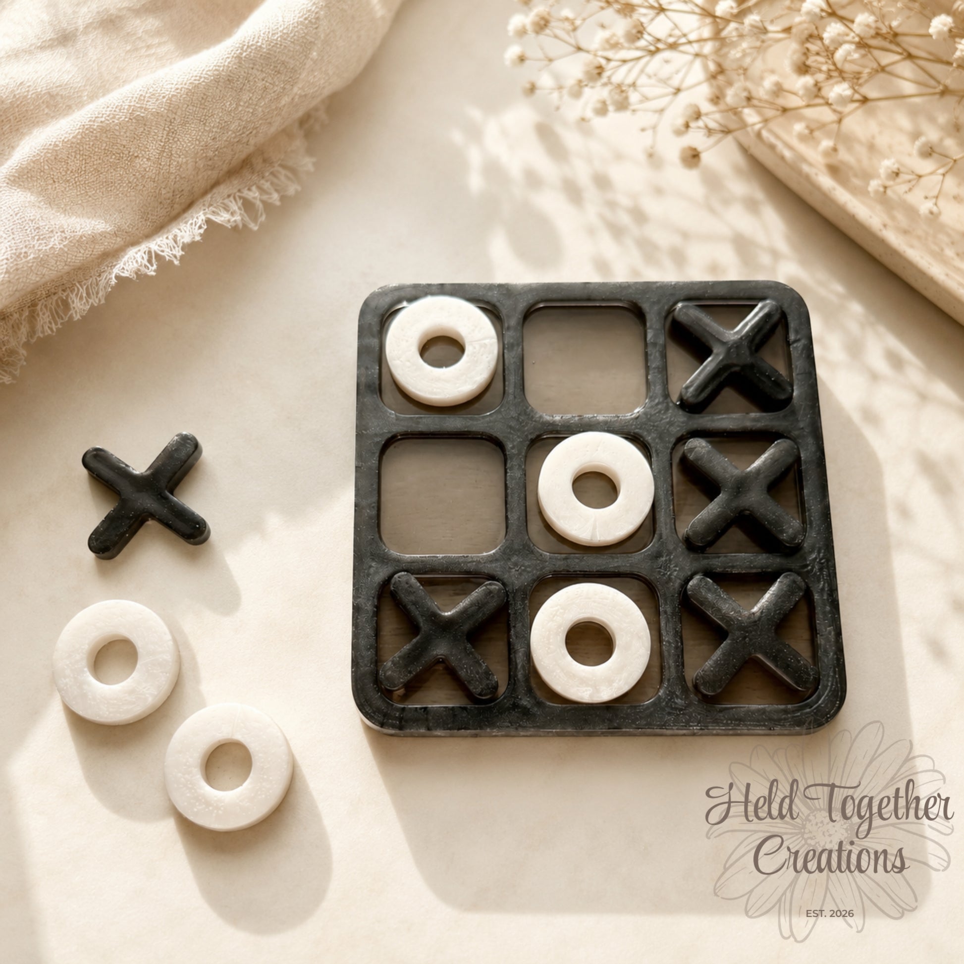 Handmade monochrome resin tic-tac-toe game with black board, black X pieces and white O pieces on a soft neutral background.