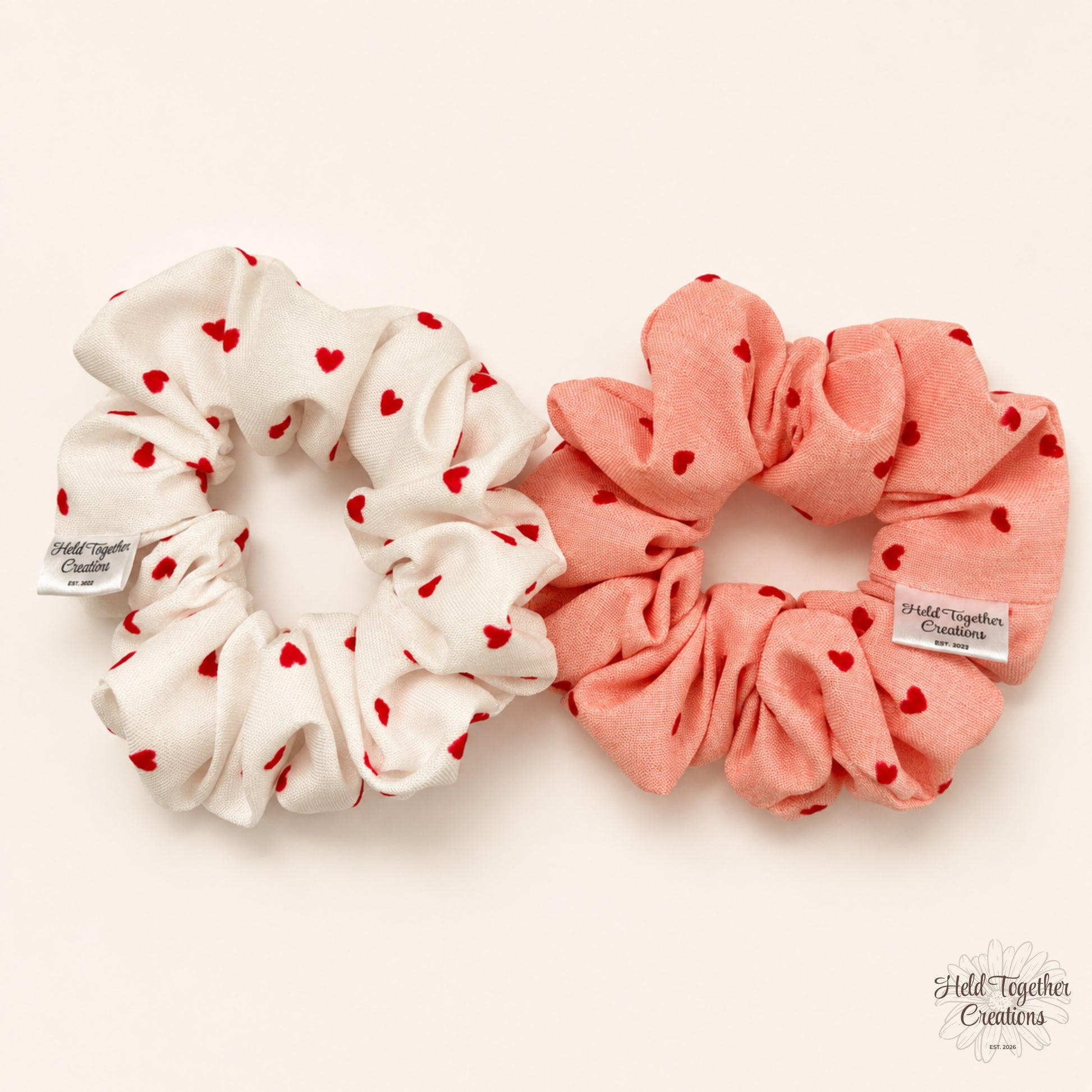 Two fabric hair scrunchies with small red heart prints, one in white and one in soft coral pink, displayed on a light cream background. Both have visible 'Held Together Creations' brand tags.