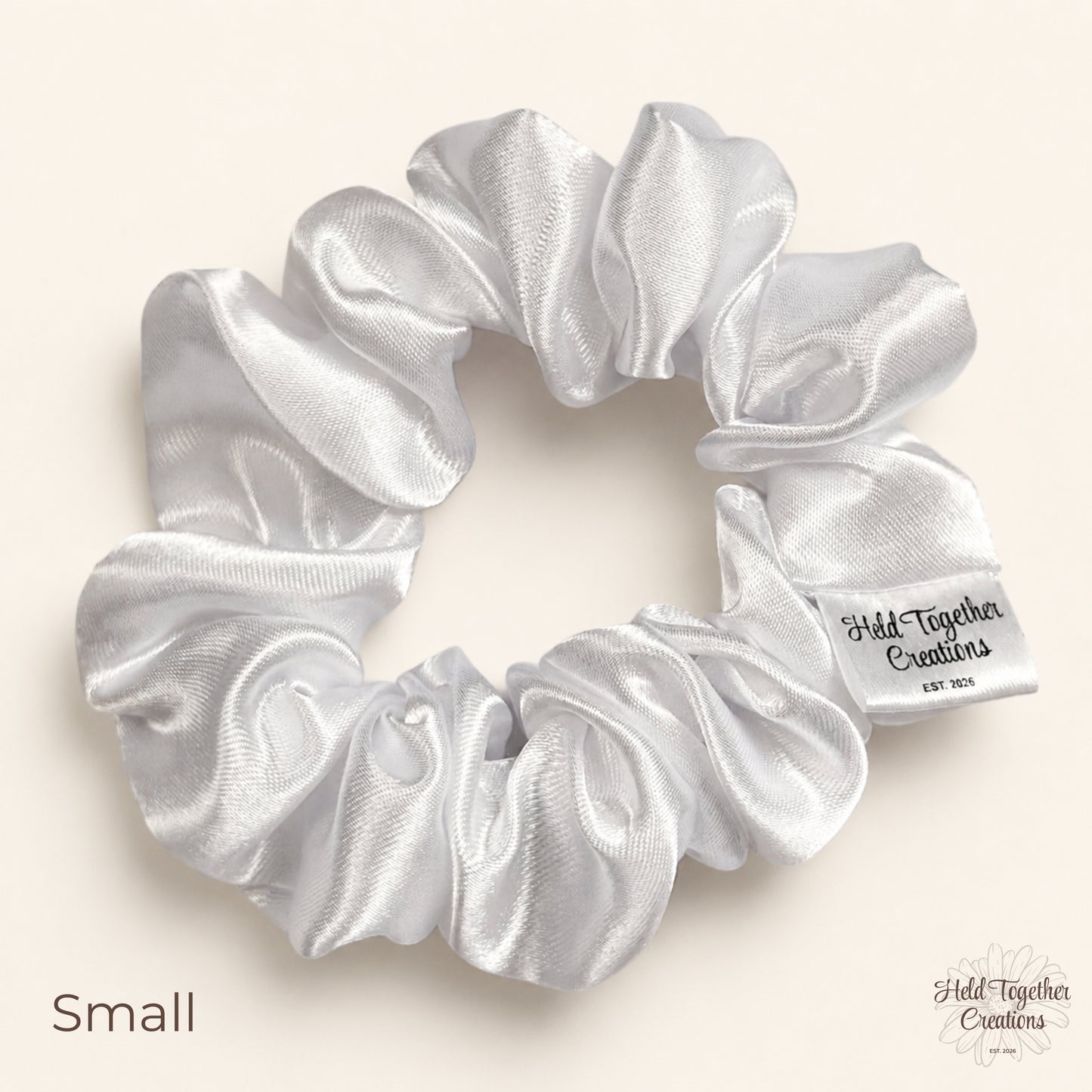 Small white satin scrunchie on a warm neutral background with Held Together Creations label visible.