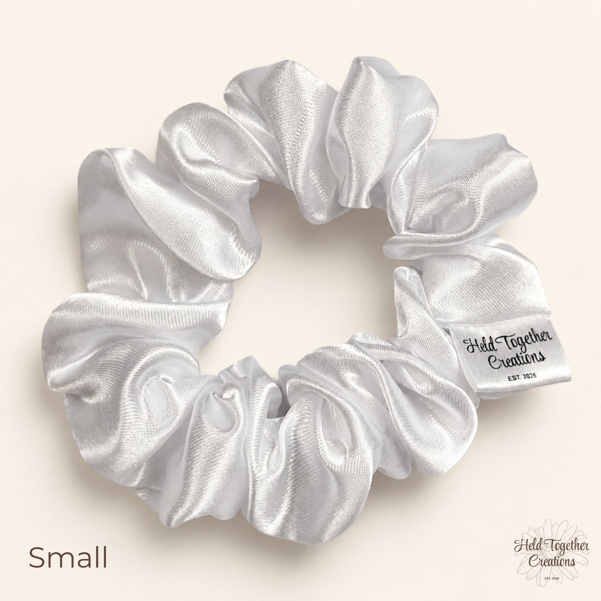 Small white satin scrunchie on a warm neutral background with Held Together Creations label visible.