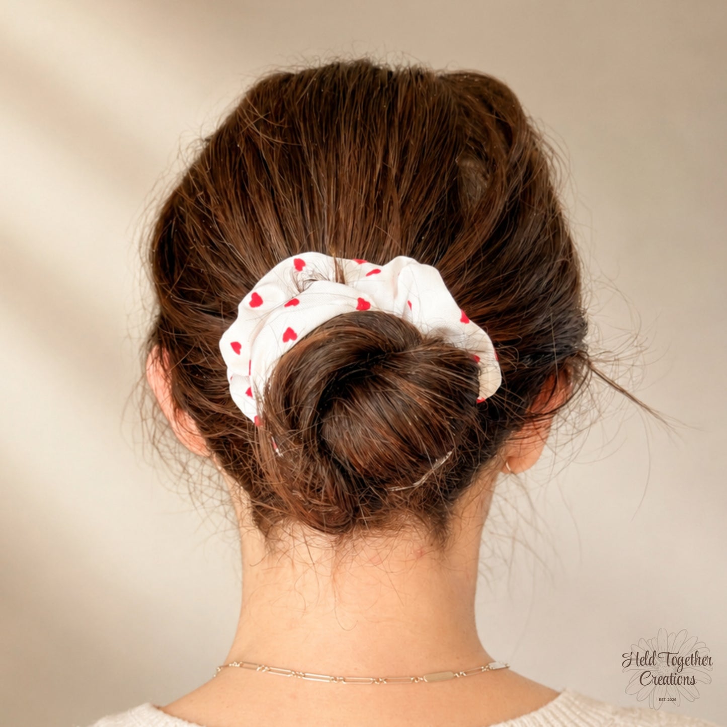 A white fabric scrunchie with small red hearts, shown on a model's brown hair in a low bun, photographed from the back against a warm beige background.