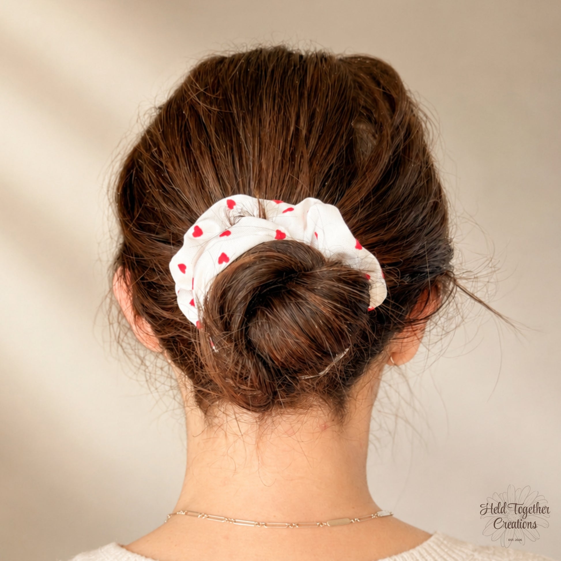 A white fabric scrunchie with small red hearts, shown on a model's brown hair in a low bun, photographed from the back against a warm beige background.