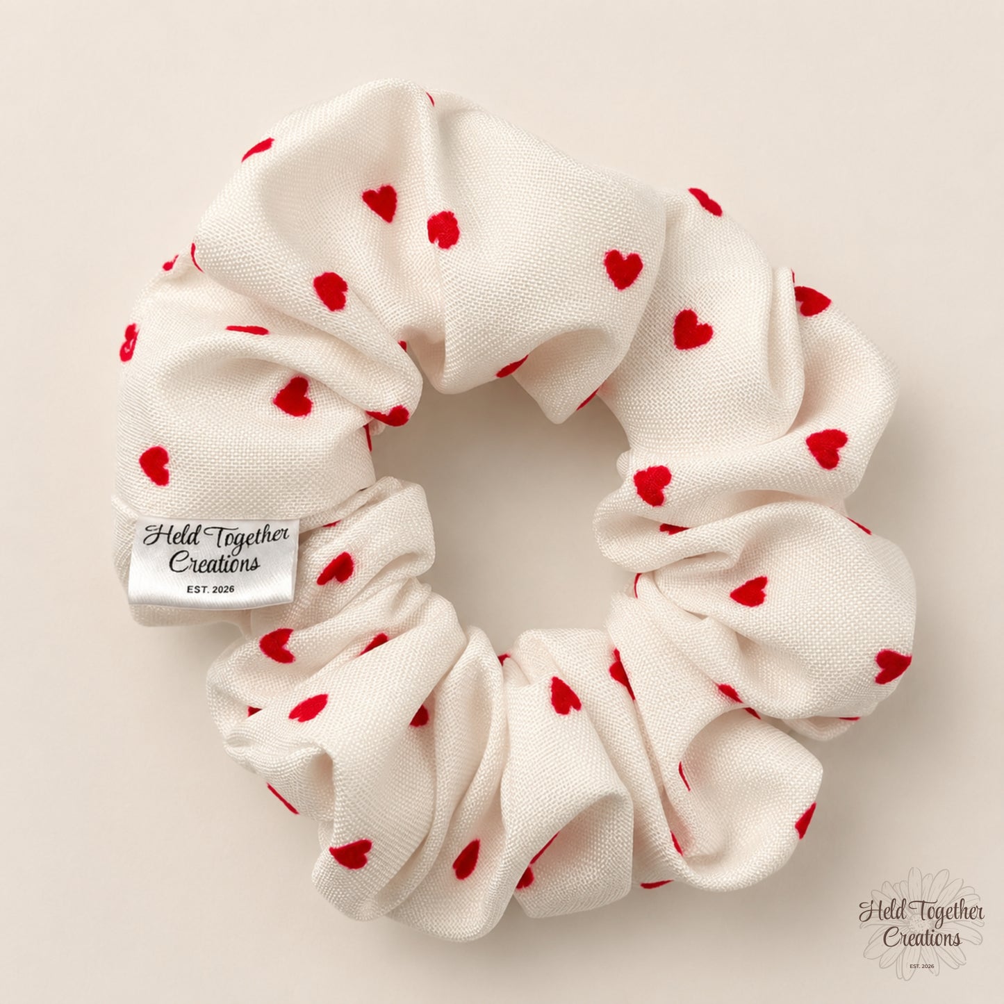 An off-white fabric hair scrunchie with small red heart prints, featuring a silver metal brand tag that reads 'Held Together Creations'. The scrunchie is photographed flat against a solid, warm beige background.