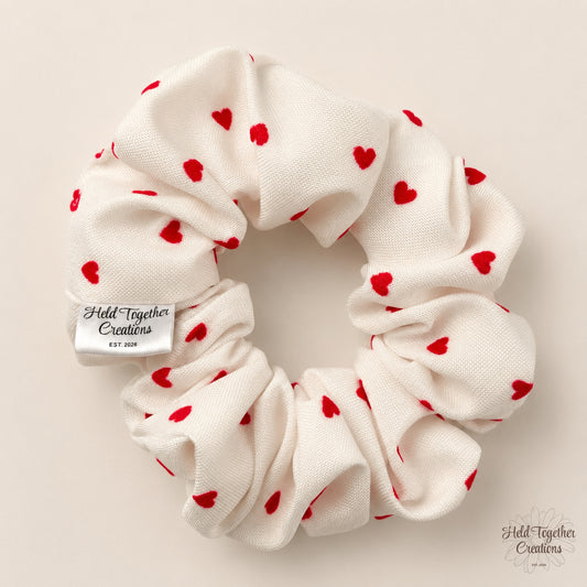 An off-white fabric hair scrunchie with small red heart prints, featuring a silver metal brand tag that reads 'Held Together Creations'. The scrunchie is photographed flat against a solid, warm beige background.