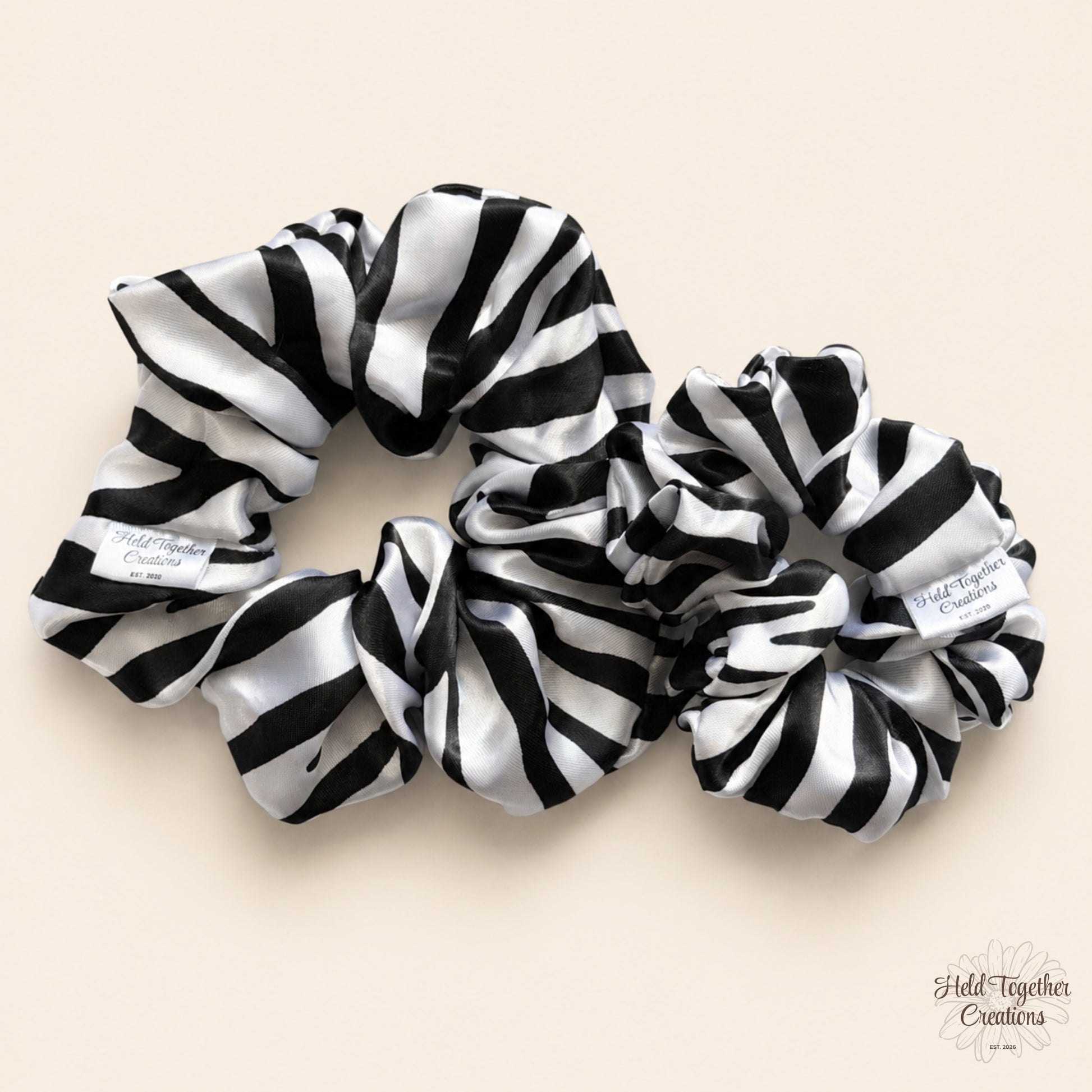 Two black and white striped satin hair scrunchies on a light beige background, with a small brand tag visible on each scrunchie.